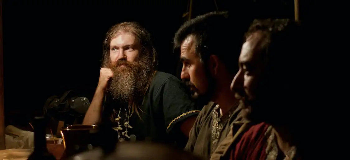 A group of Viking men sit at a table, wearing traditional dress. Viking shopping can source items like the Viking jewelry seen on one of the Vikings