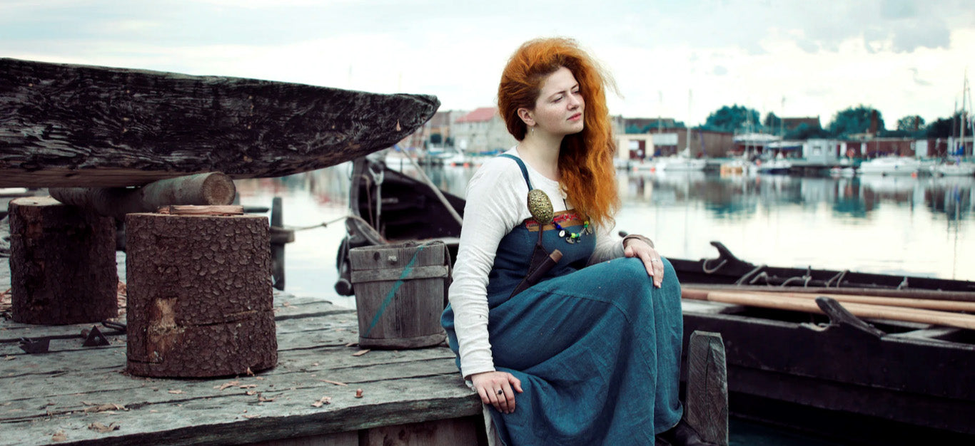 A red haired woman sits on a dock near a longship, looking out to sea, wearing a Viking inspired outfit with Norse jewelry