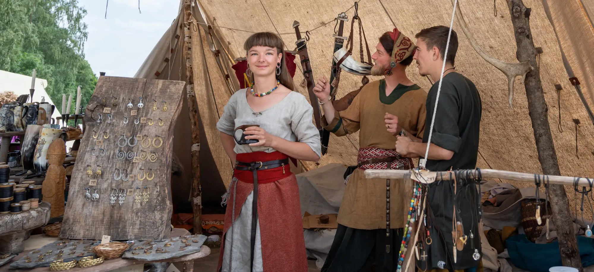 A modern day female Viking enthusiast stands wearing Viking dress in a Norse themed village. She is standing next to a woman Viking jewelry display stand