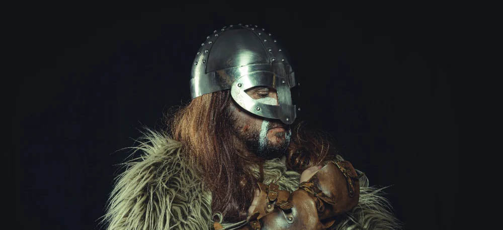A man dressed as a Viking stands against a black background, looking off to the side. He wears leather gloves and a Viking helmet.