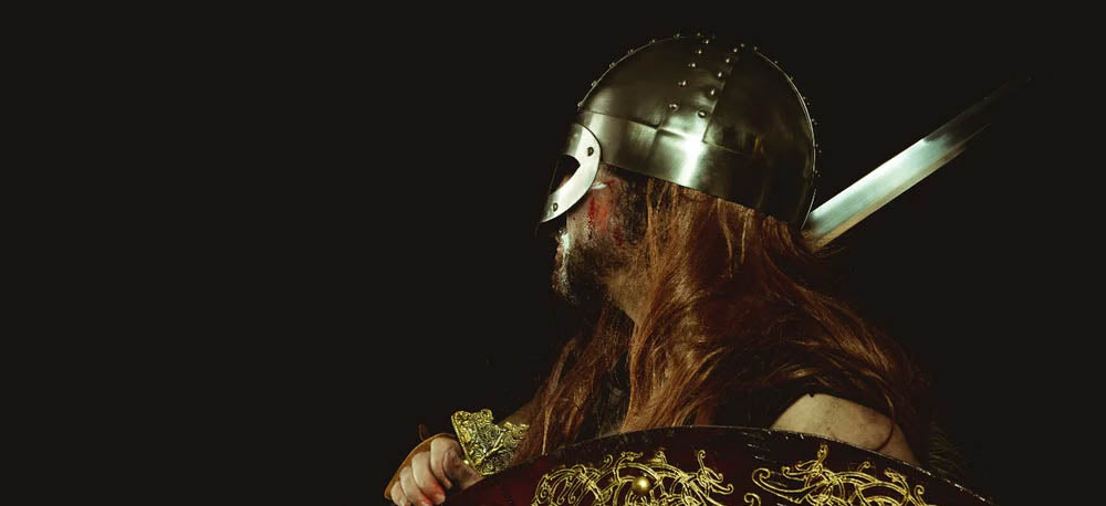 A man dressed as a Viking stands holding a sword facing away from the camera. He holds a shield in his left hand