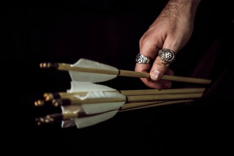 A mans hand is in frame, wearing Viking style rings and picking up an arrow from a set 