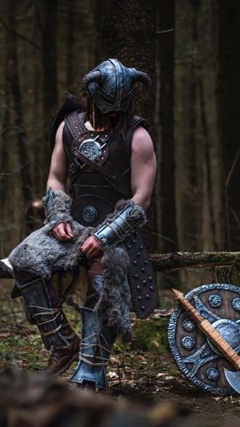 A man dressed in a Viking inspired outfit sits on a tree branch in the woods, with a Norse themed shield and sword by his side 